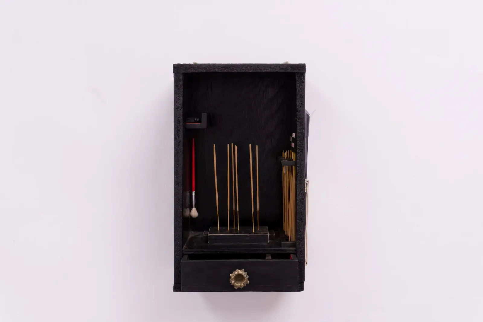 A small black wooden shrine mounted on a wall, holding incense sticks, a brush and a lighter.