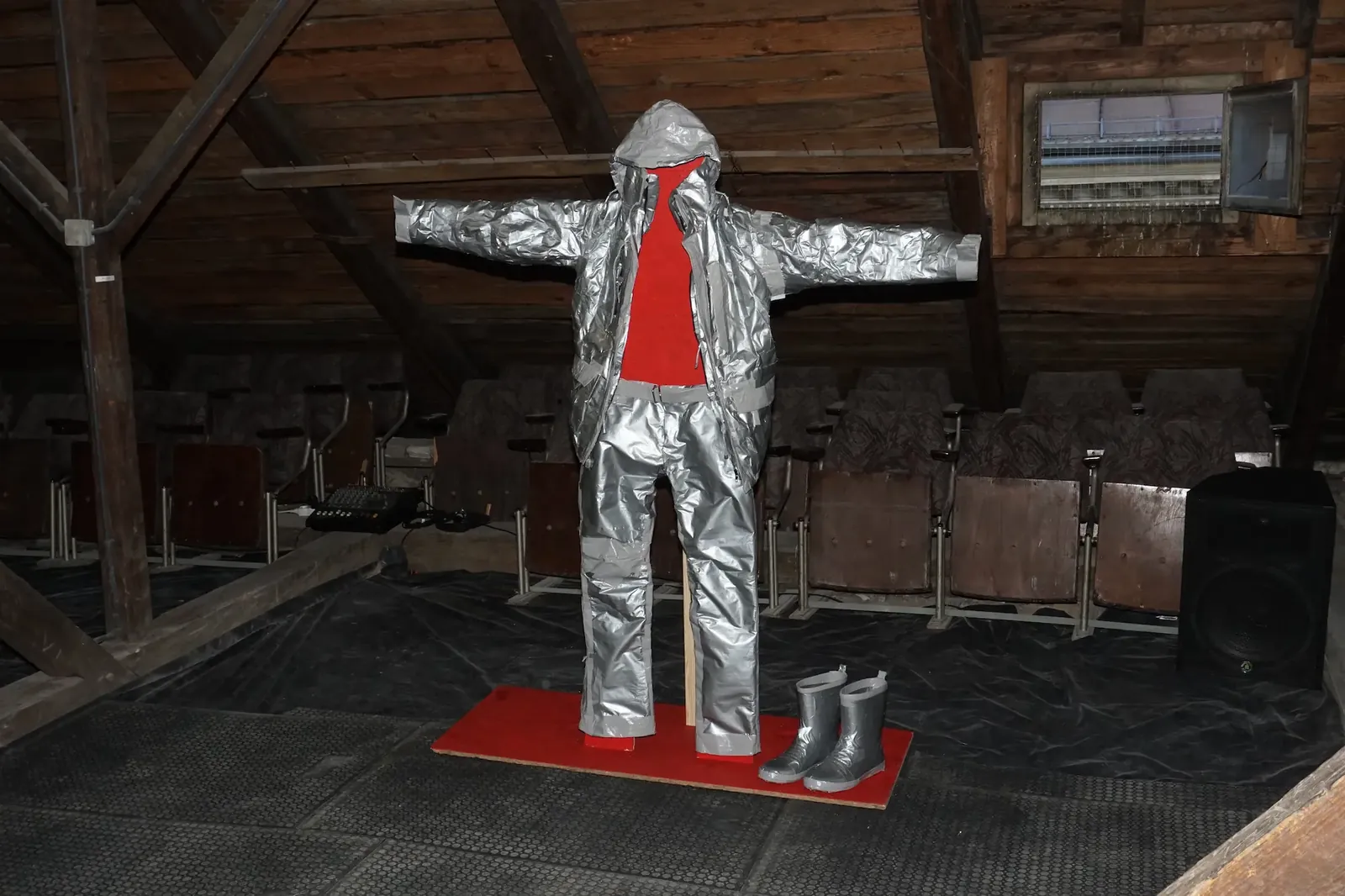 A work suit entirely wrapped in silver duct tape, standing upright in an attic space on a red mat.